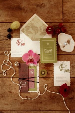Elegant wedding stationery flat lay with pearl necklace, pink orchid, and botanical envelope design on wooden surface.