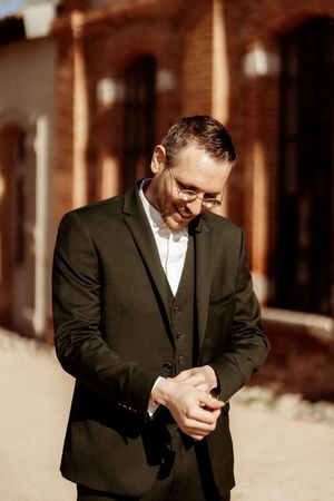 A groom in a dark suit and glasses looks down with a joyful smile while adjusting his cufflinks outdoors.