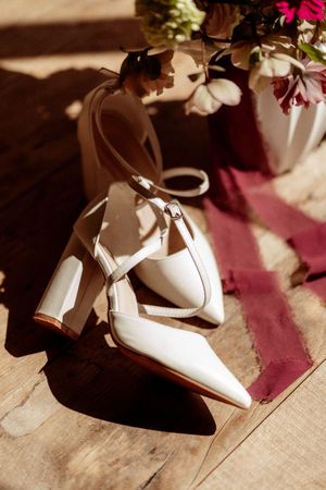 Elegant white bridal shoes with ankle straps cast shadows on wooden floor next to flowers.