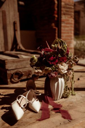 White bridal shoes and a floral arrangement with deep red and cream flowers in a vase casting dramatic shadows.
