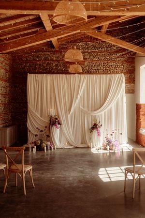 Elegant white draped backdrop with floral arrangements in a rustic brick venue with string lights and wooden beams.