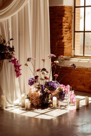 Romantic floral arrangement with purple and pink flowers next to white candles in front of window with flowing drapes.