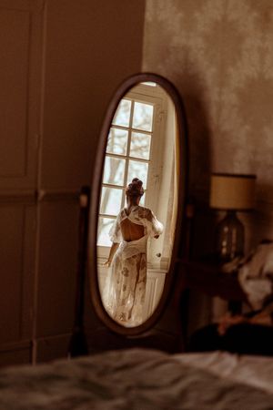 Artistic reflection in oval vintage mirror shows figure in white dress by bright window.