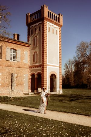 A wedding couple poses in front of a historic brick mansion with white trim and arched windows on a sunny day.
