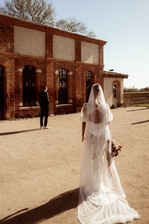 Bride in flowing white gown and veil stands behind groom for first look moment at rustic brick venue.