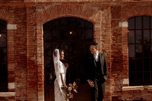 Wedding couple stands together in front of rustic brick archway with dramatic lighting.