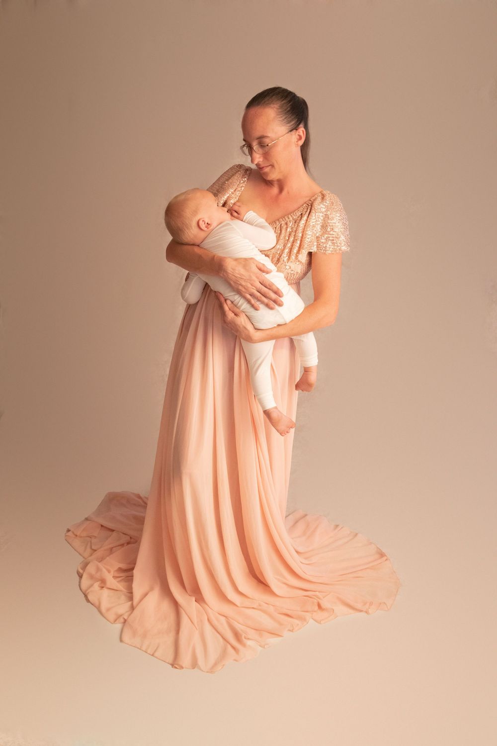 Mother in elegant peach sequin gown with long train tenderly holding sleeping newborn baby.