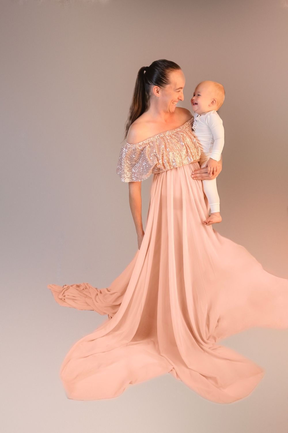 Joyful mother in off-shoulder peach sequin gown holding laughing baby against neutral backdrop.