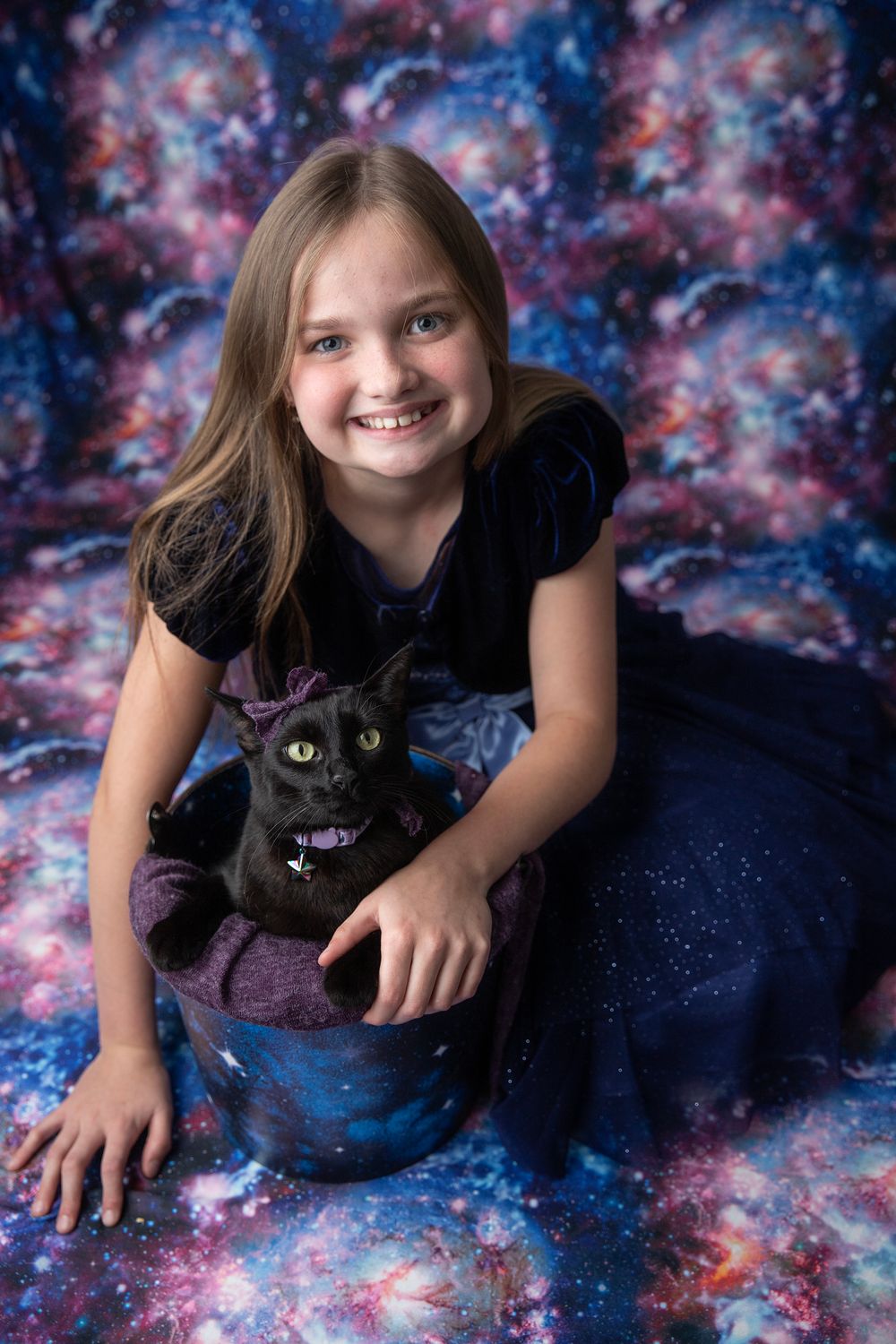 Smiling girl in navy dress holding black cat in bucket against colorful galaxy-themed backdrop.