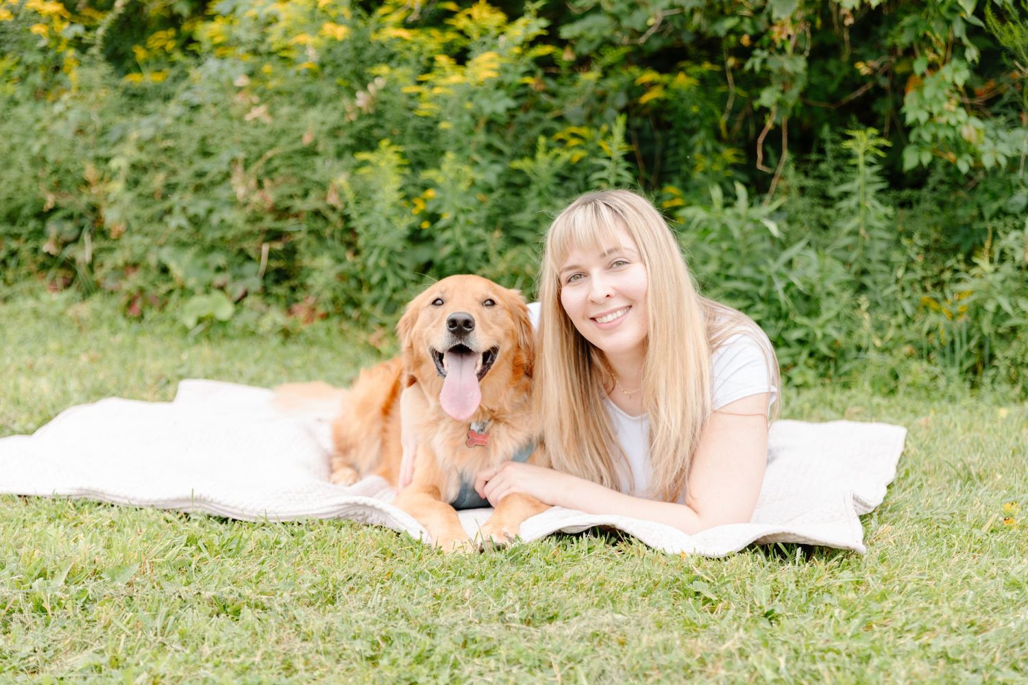Woman lying on blanket outdoors with golden retriever dog amid greenery.