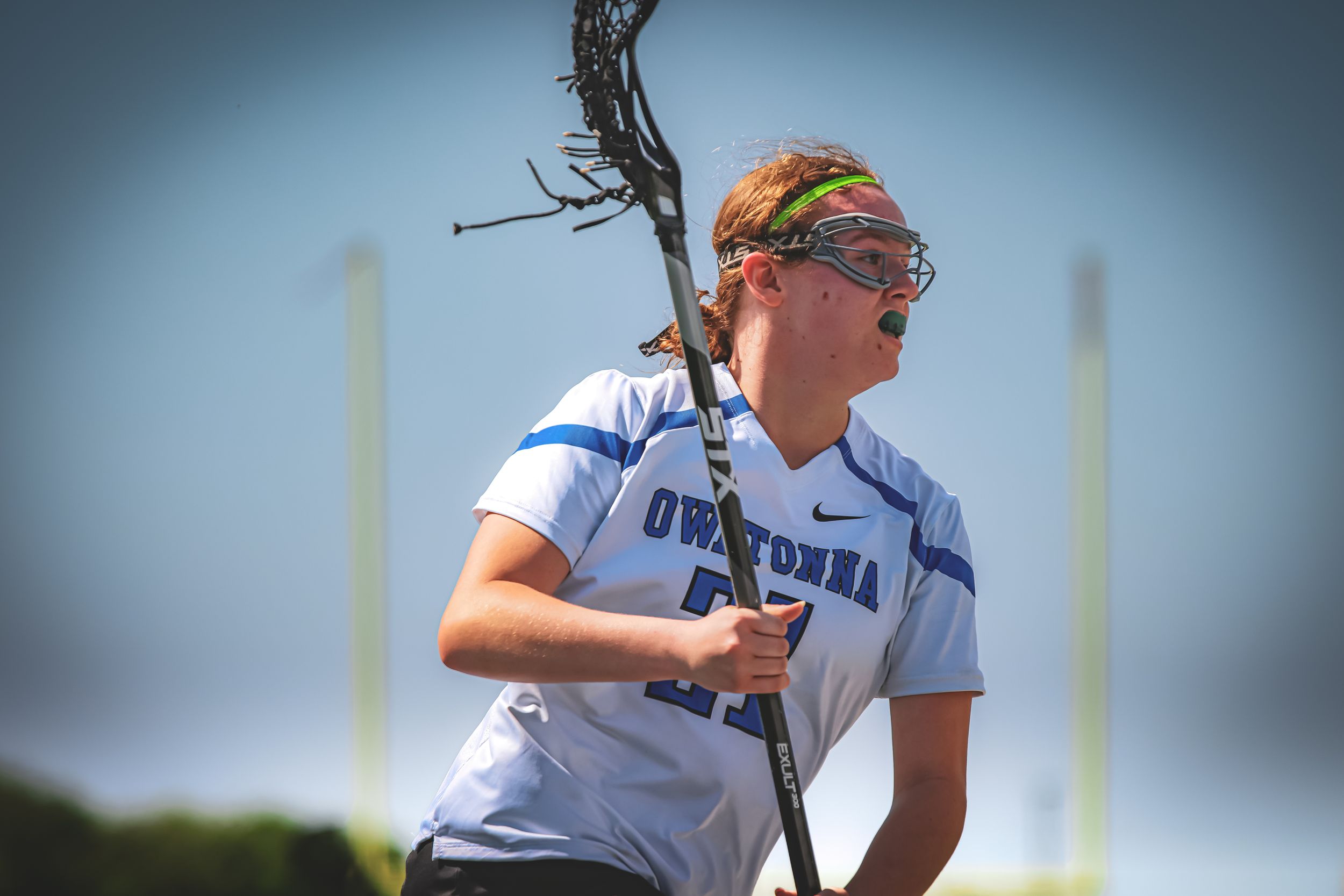 A lacrosse player in white jersey and colorful headband holds their stick during an outdoor game.