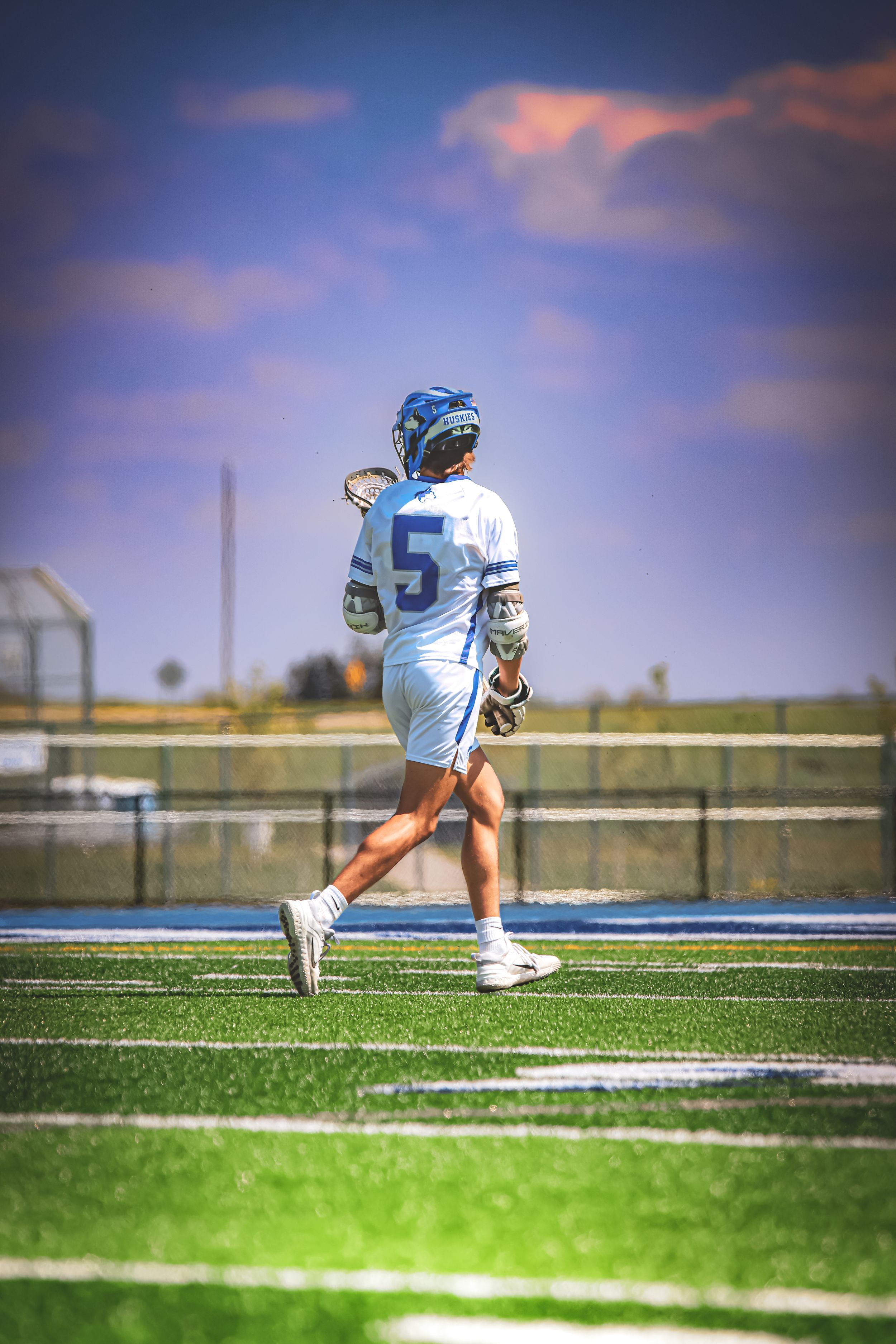 A lacrosse player in white uniform number 5 runs across a turf field under colorful sunset skies.