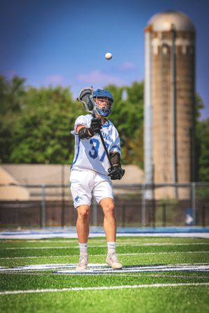 A lacrosse player throws a ball with a large silo visible in the background.