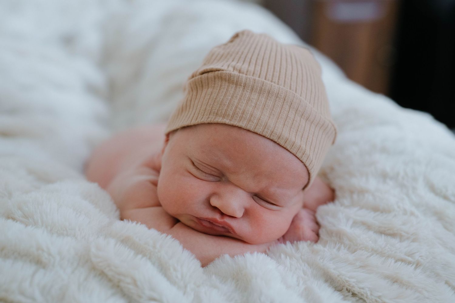 Newborn baby sleeping peacefully on soft white blanket wearing beige knit hat.