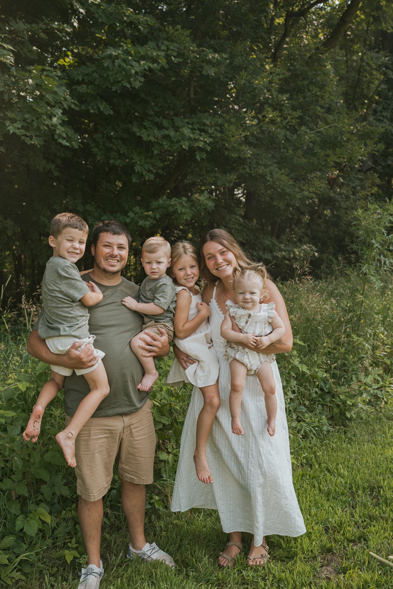 Family group poses together outdoors in coordinated casual outfits against lush green foliage background.