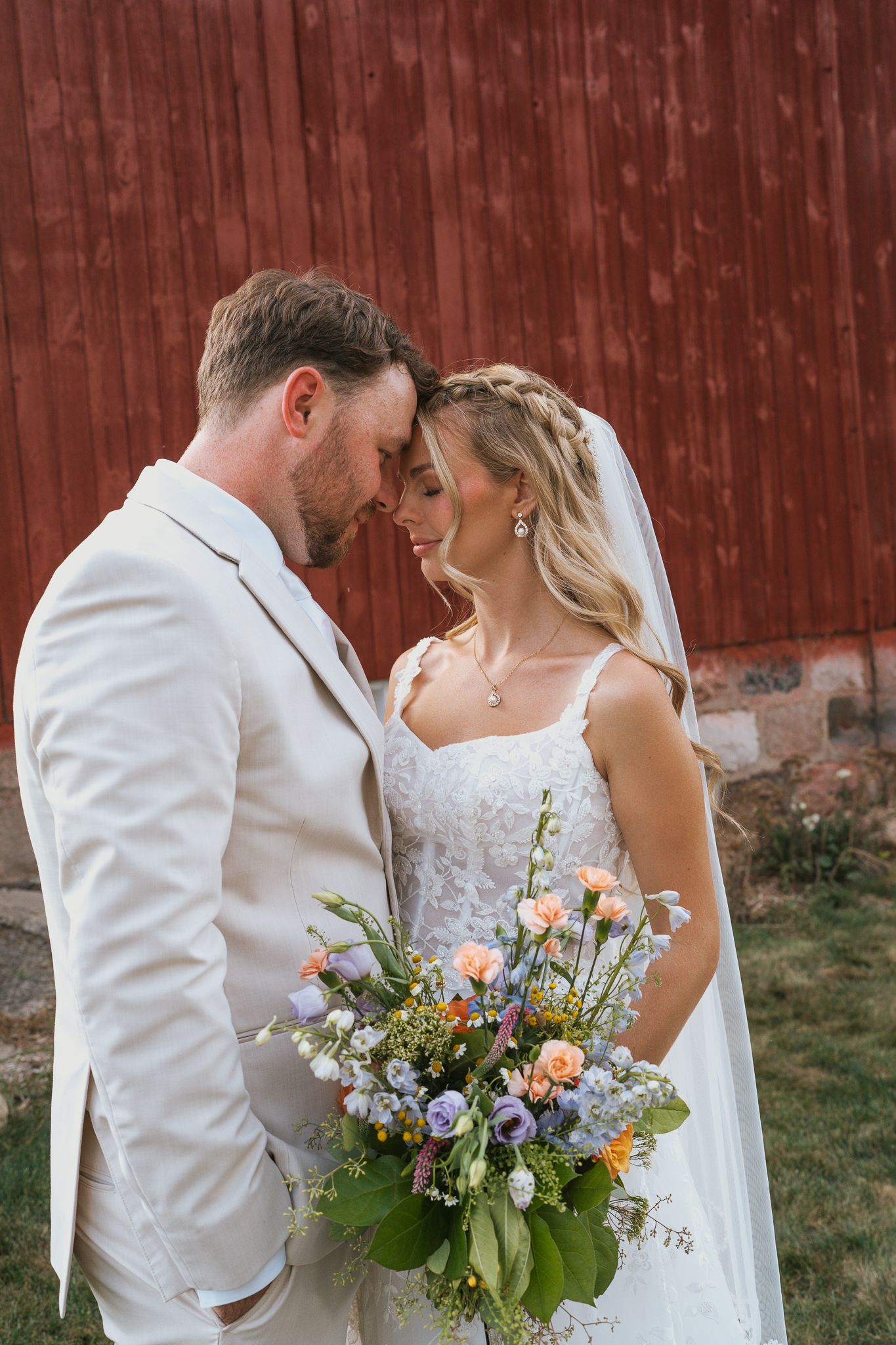 Couple in wedding attire sharing romantic moments against a rustic red barn wall with wildflower bouquet.