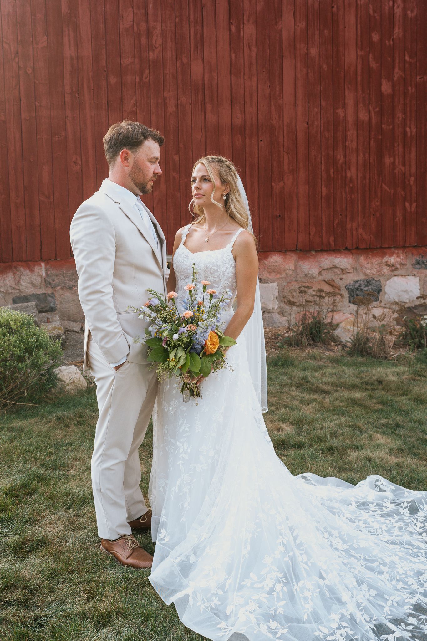 A couple in wedding attire stands against a rustic red barn wall as the bride's lace dress trails elegantly behind them.