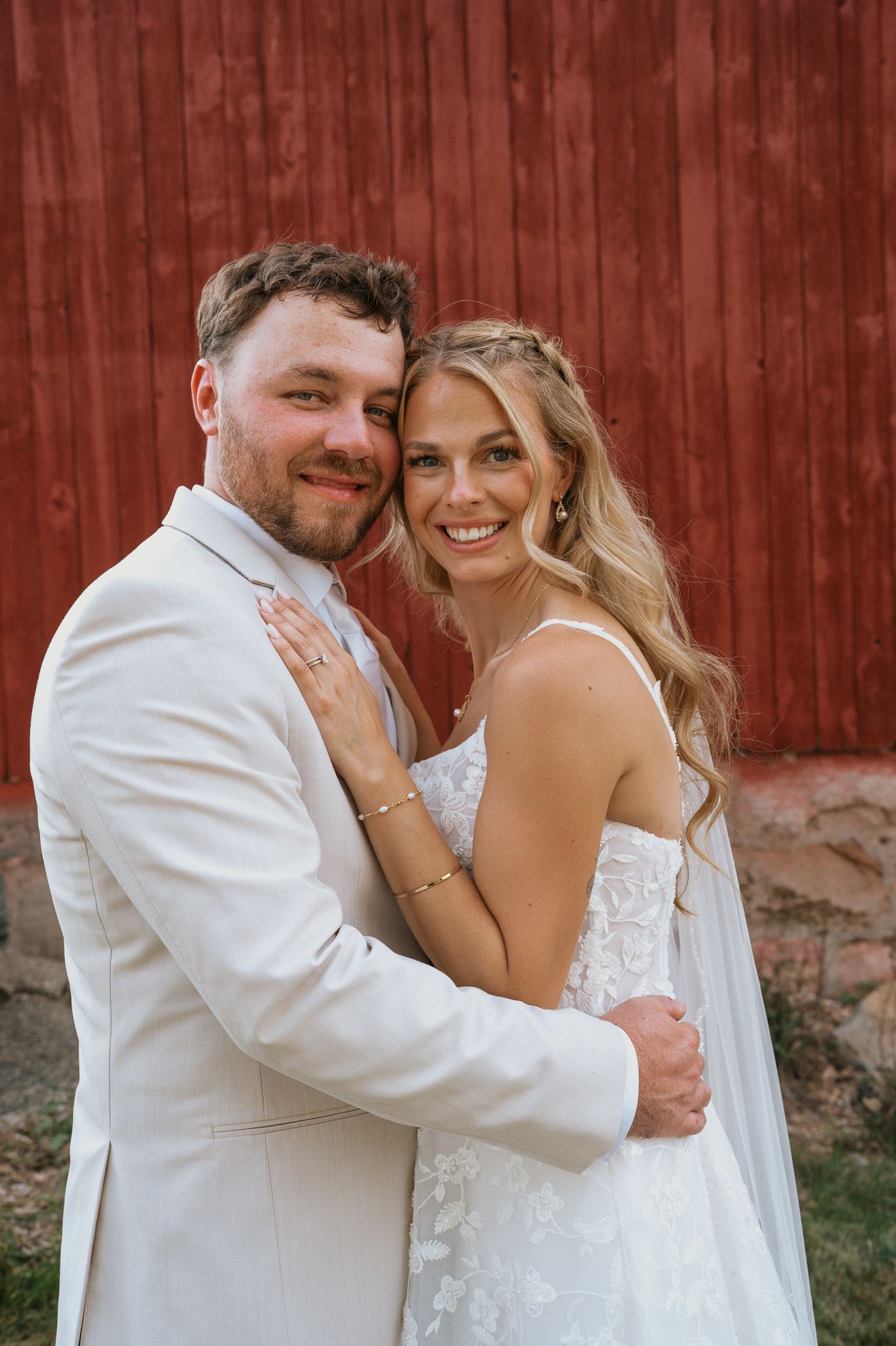 A couple in wedding attire share intimate moments against a rustic red barn backdrop in a series of candid photos.