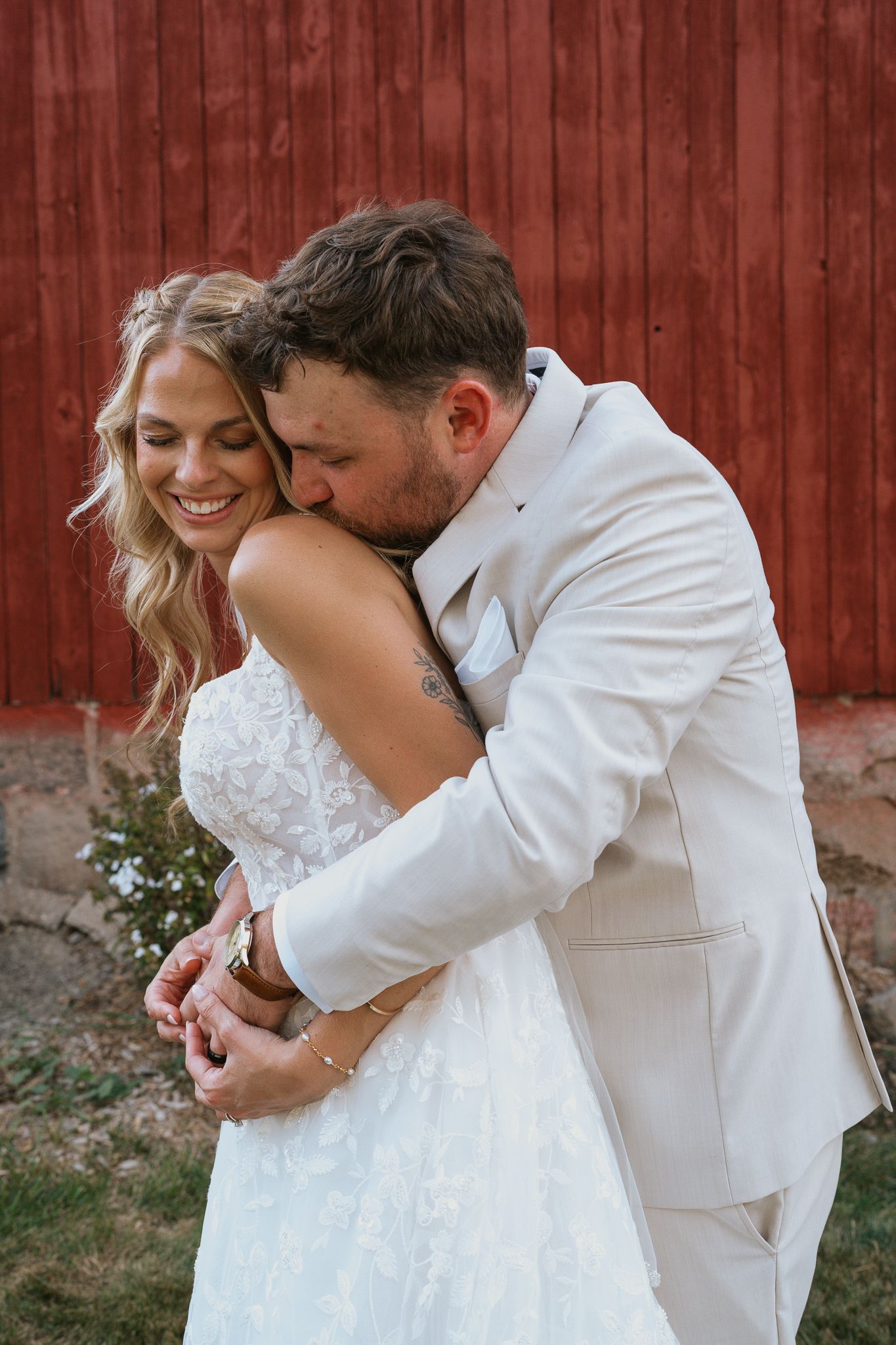A romantic wedding couple in white embraces and dances together against a rustic red barn wall.
