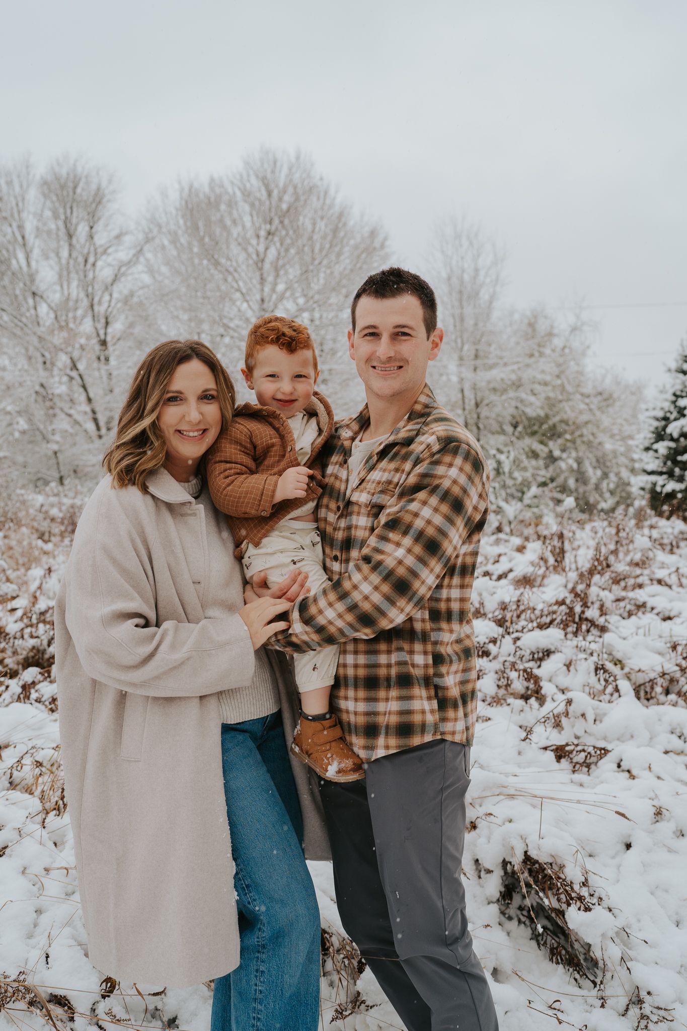 A family poses together in a snowy winter landscape wearing cozy sweaters and plaid.