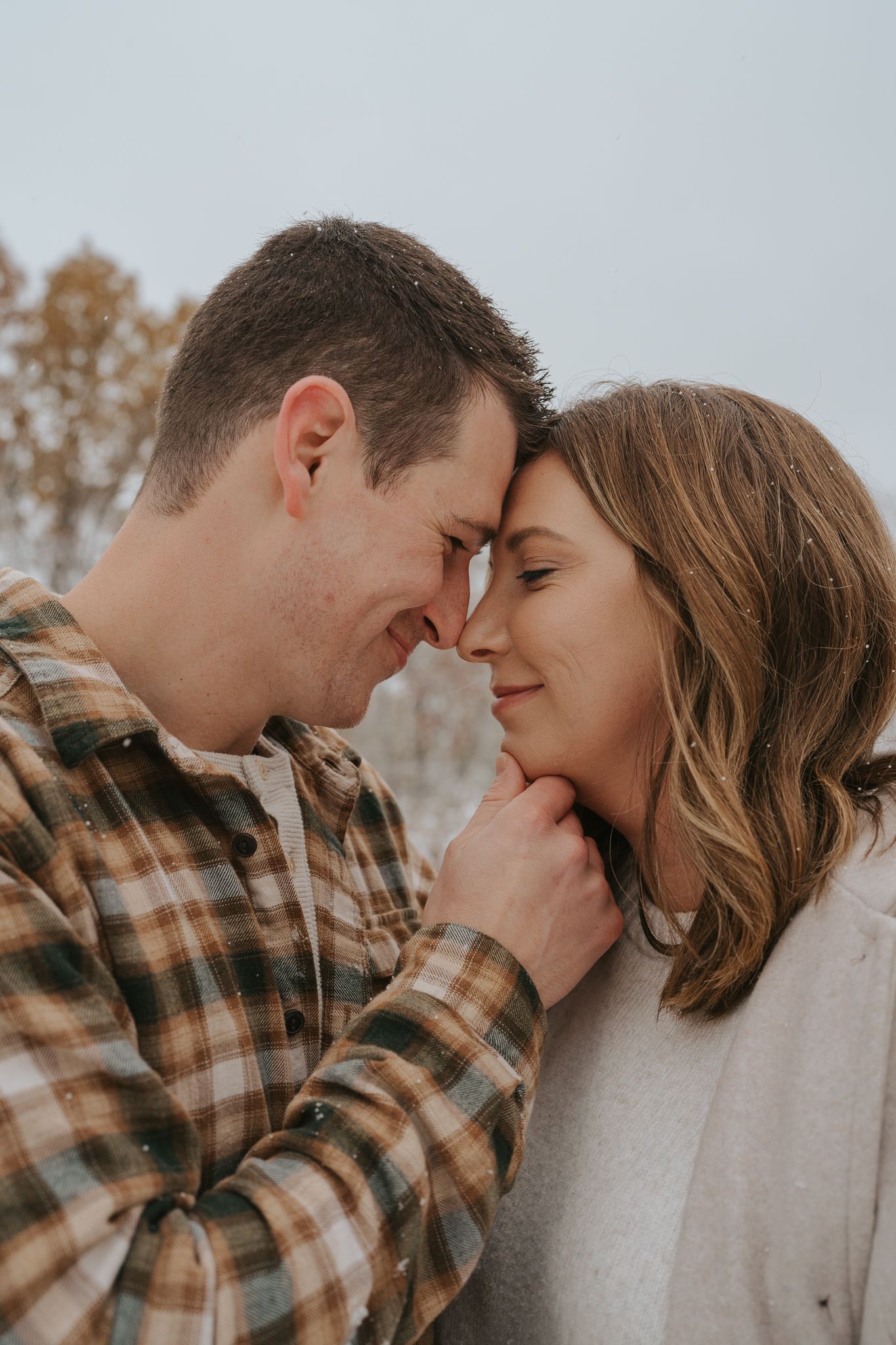 A romantic couple shares an intimate moment outdoors, touching foreheads while wearing casual fall attire.