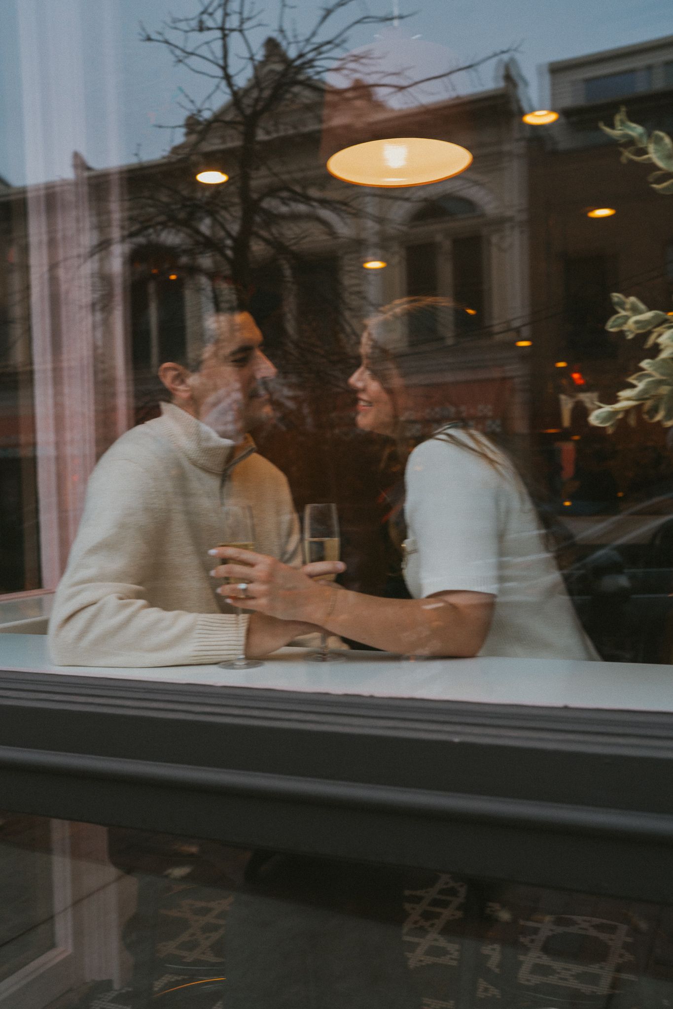 A couple shares drinks and conversation through a cafe window at dusk with warm lighting overhead.