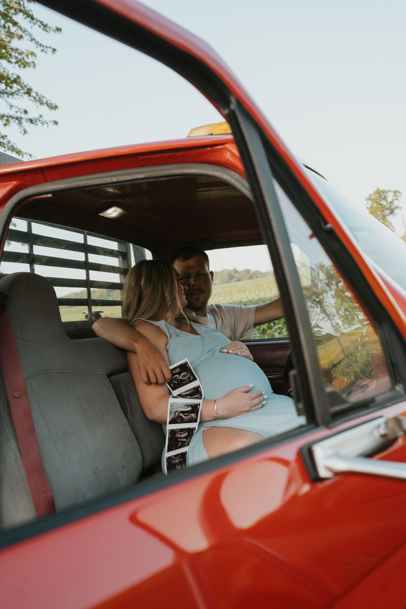 A couple sits in a vintage red truck with ultrasound photos, capturing a tender pregnancy announcement moment.