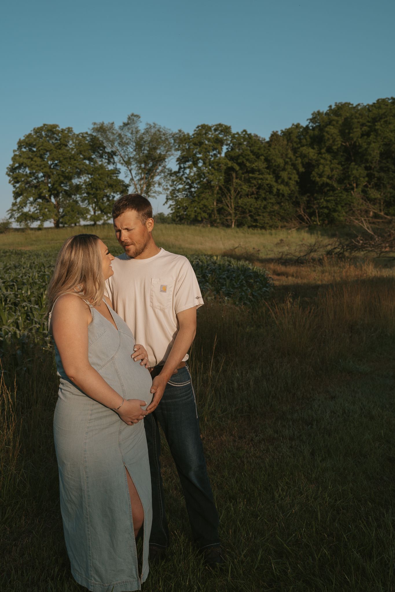 A romantic maternity photoshoot in a sunlit field with a couple sharing an intimate moment at sunset.