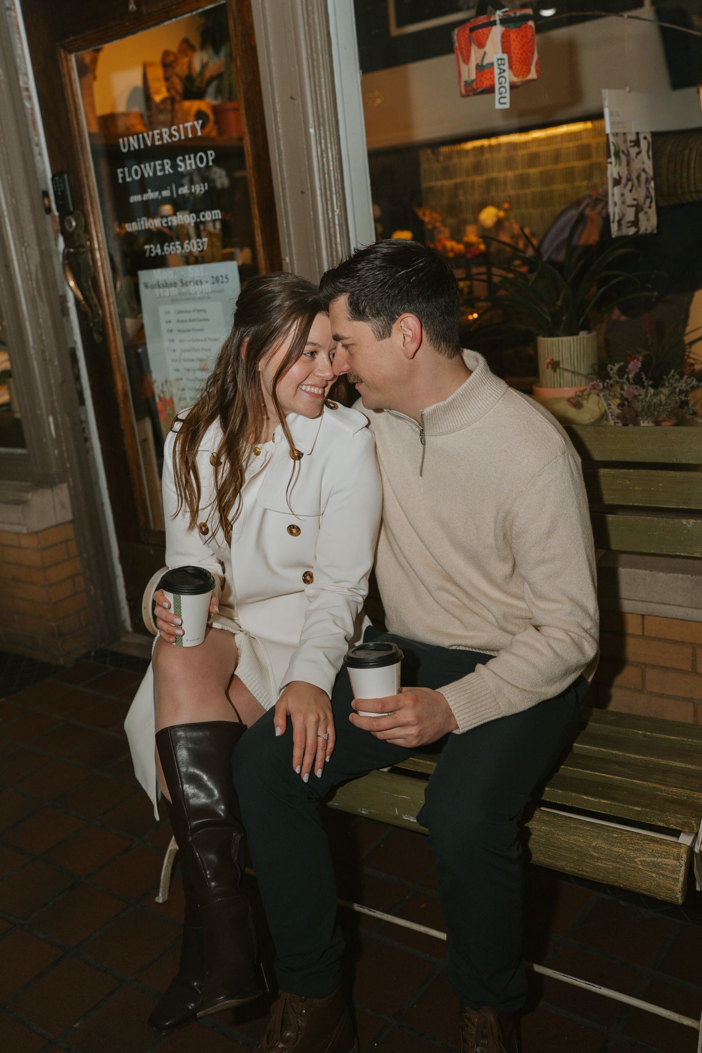 A romantic couple shares tender moments while sitting on a bench outside a storefront in the evening.