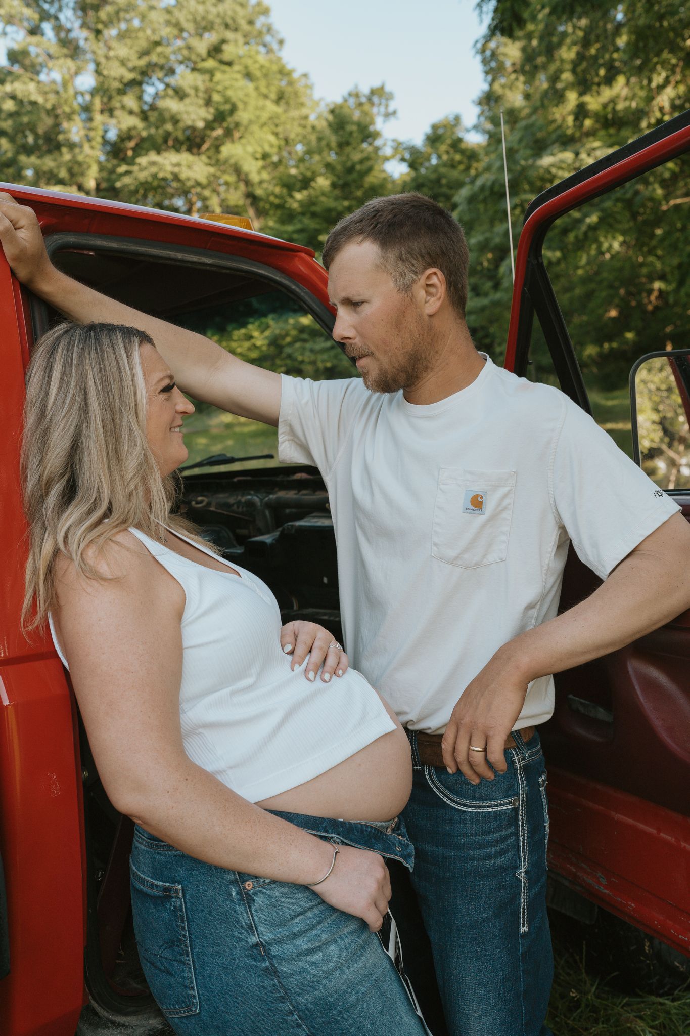 Couple in white and denim poses playfully by a red vintage truck in a sunlit outdoor setting.