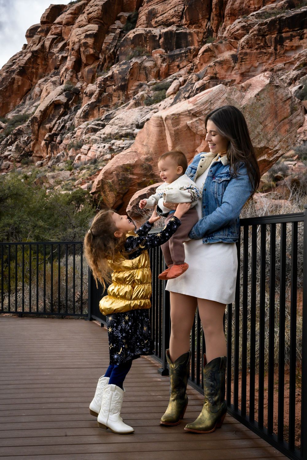 A mother holds her baby on a wooden deck at Red Rock Canyon, as her young daughter in a gold jacket looks up adoringly.