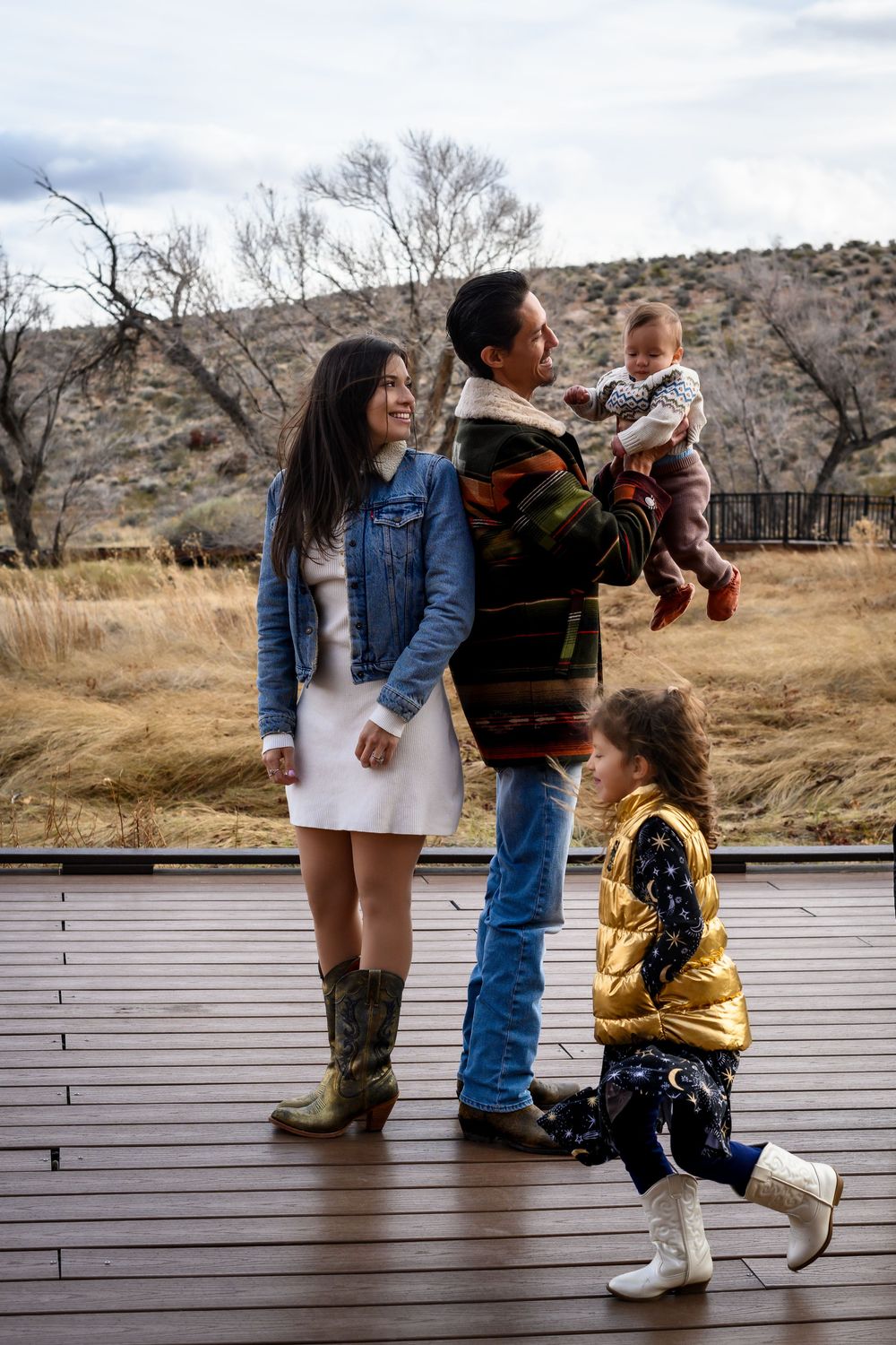 Family of four posing on wooden deck in autumn landscape, parents holding baby and toddler in gold vest.