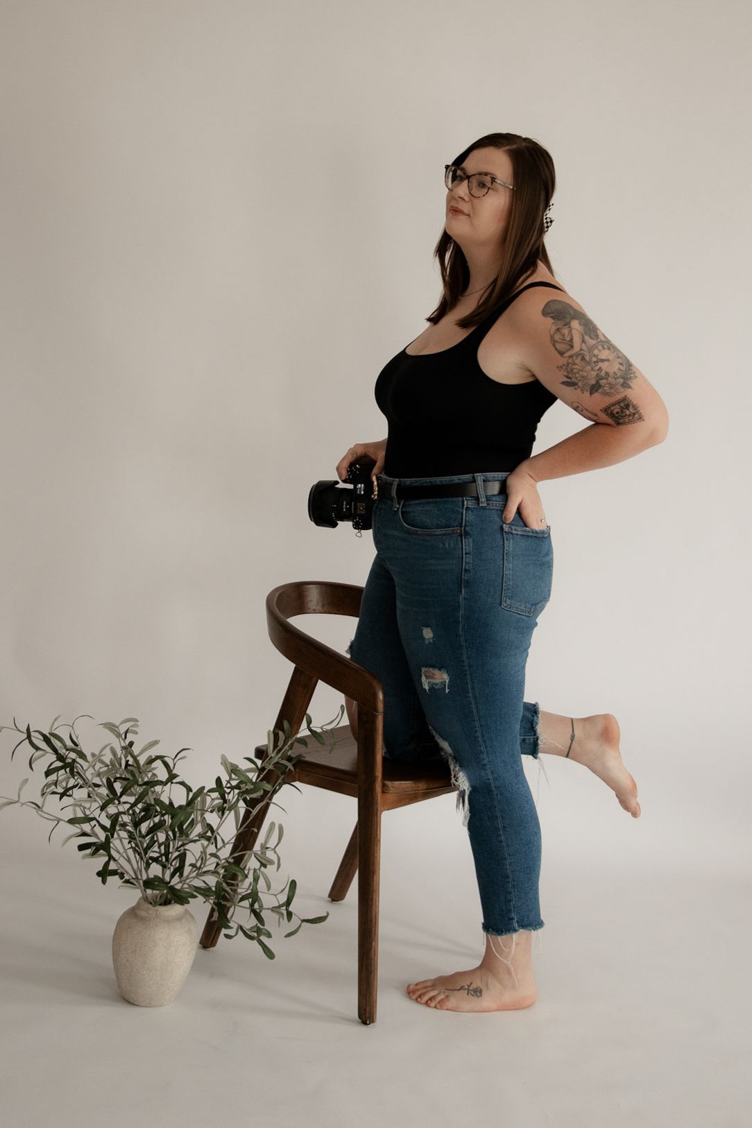 Female photographer in black tank top and ripped jeans poses with DSLR camera beside wooden chair and potted plant.