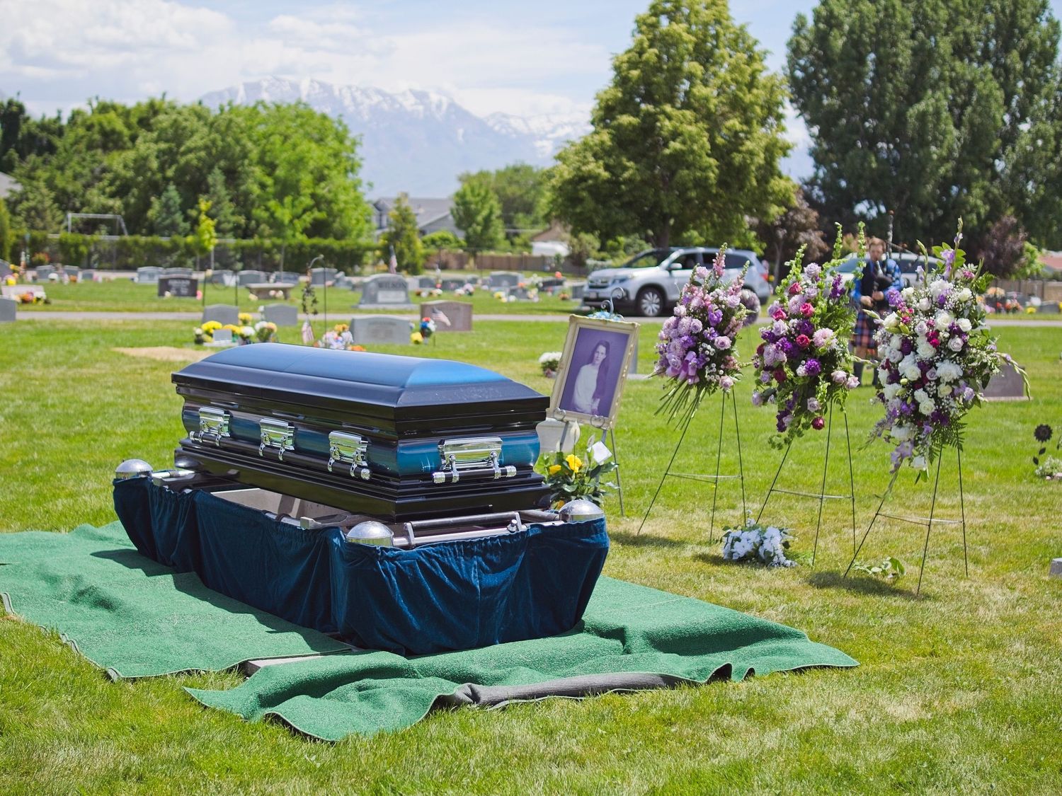 A deep blue casket rests above the gravesite. Three large flower arrangements are displayed behind the casket.