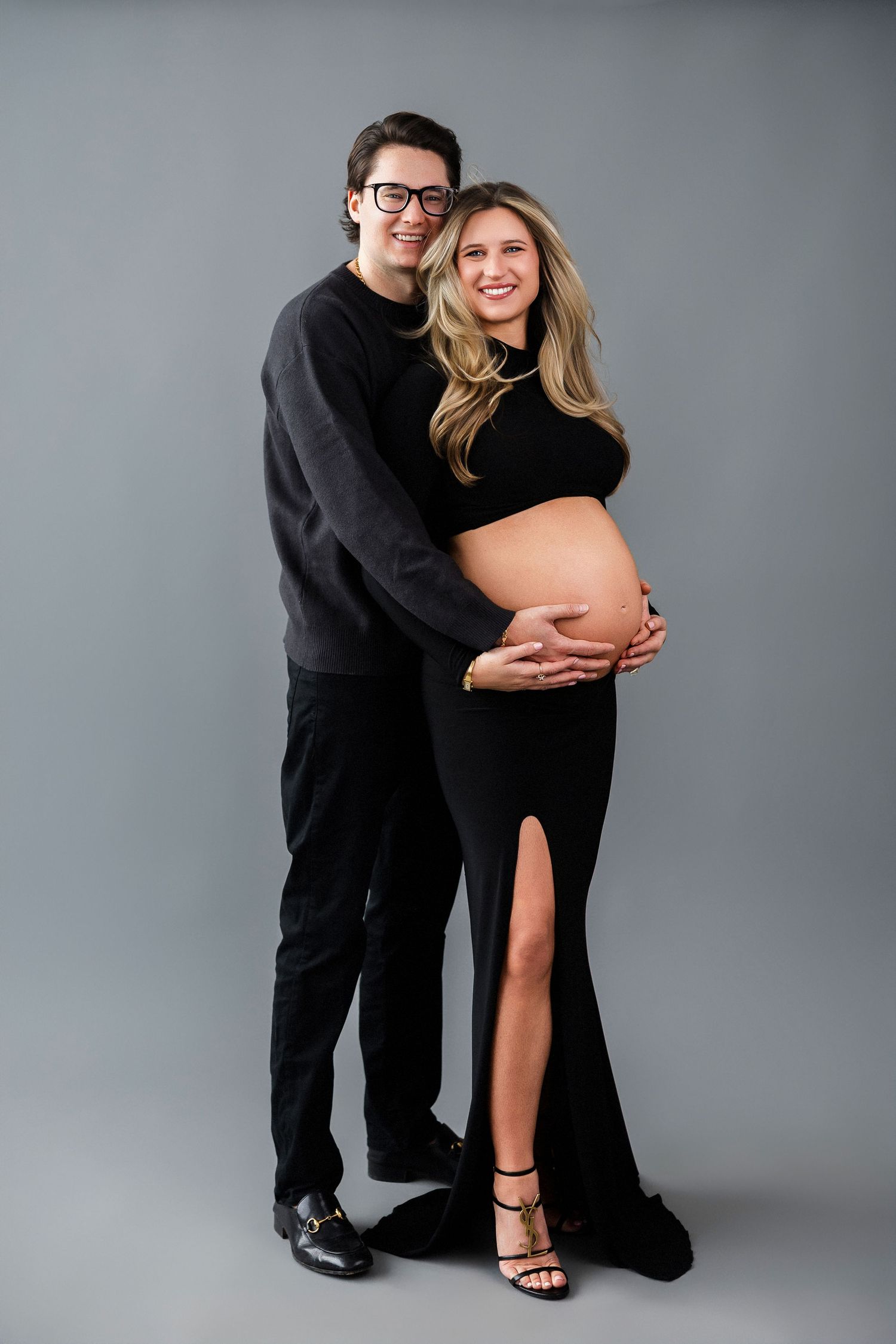 Intimate maternity portrait of couple in black attire against gray studio backdrop.