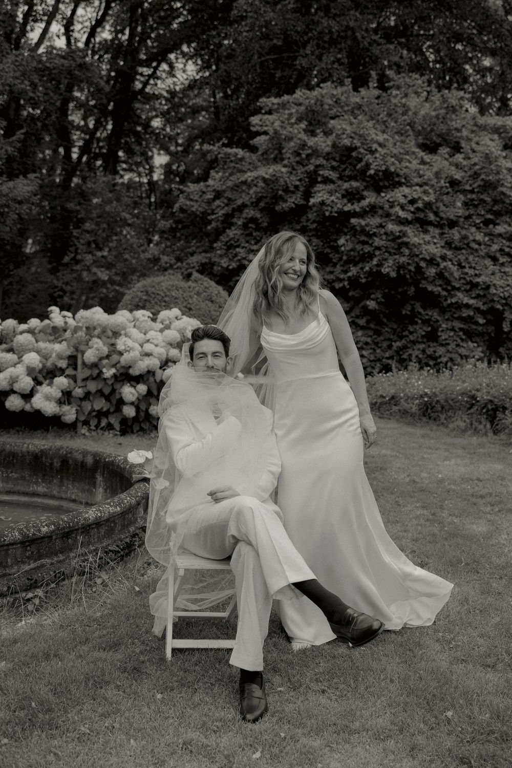 Black and white wedding portrait of a bride in a satin gown and veil standing beside a seated groom in a garden with hydrangeas.