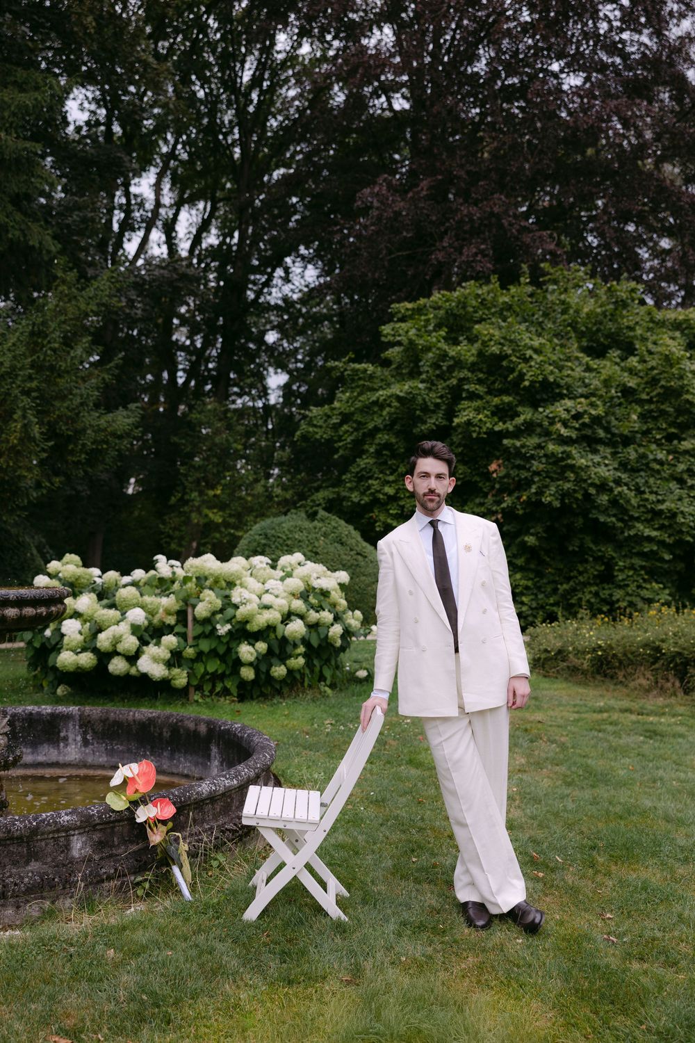 Man in a white suit and dark tie stands beside a folding chair in a lush garden with white hydrangeas and a stone fountain.