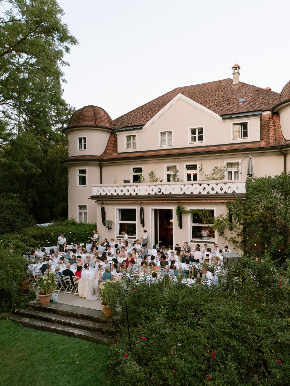 Wedding guests gather at an outdoor reception on a terrace beside an elegant cream villa with a terracotta roof and lush garden.