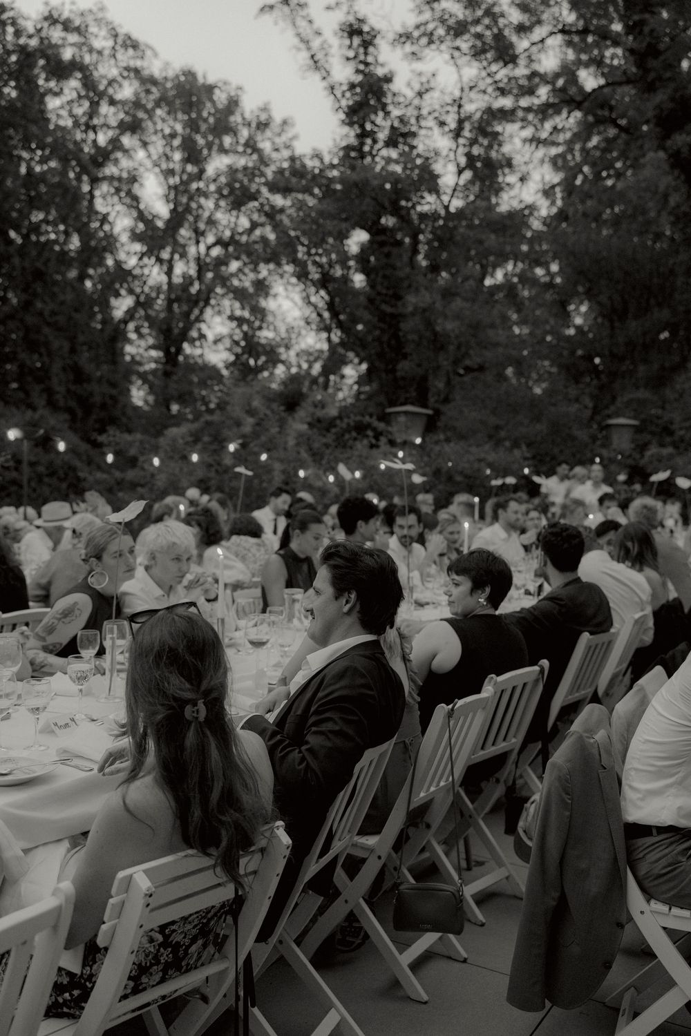 Black and white photo of wedding guests seated at long outdoor tables under string lights and tall trees at dusk.