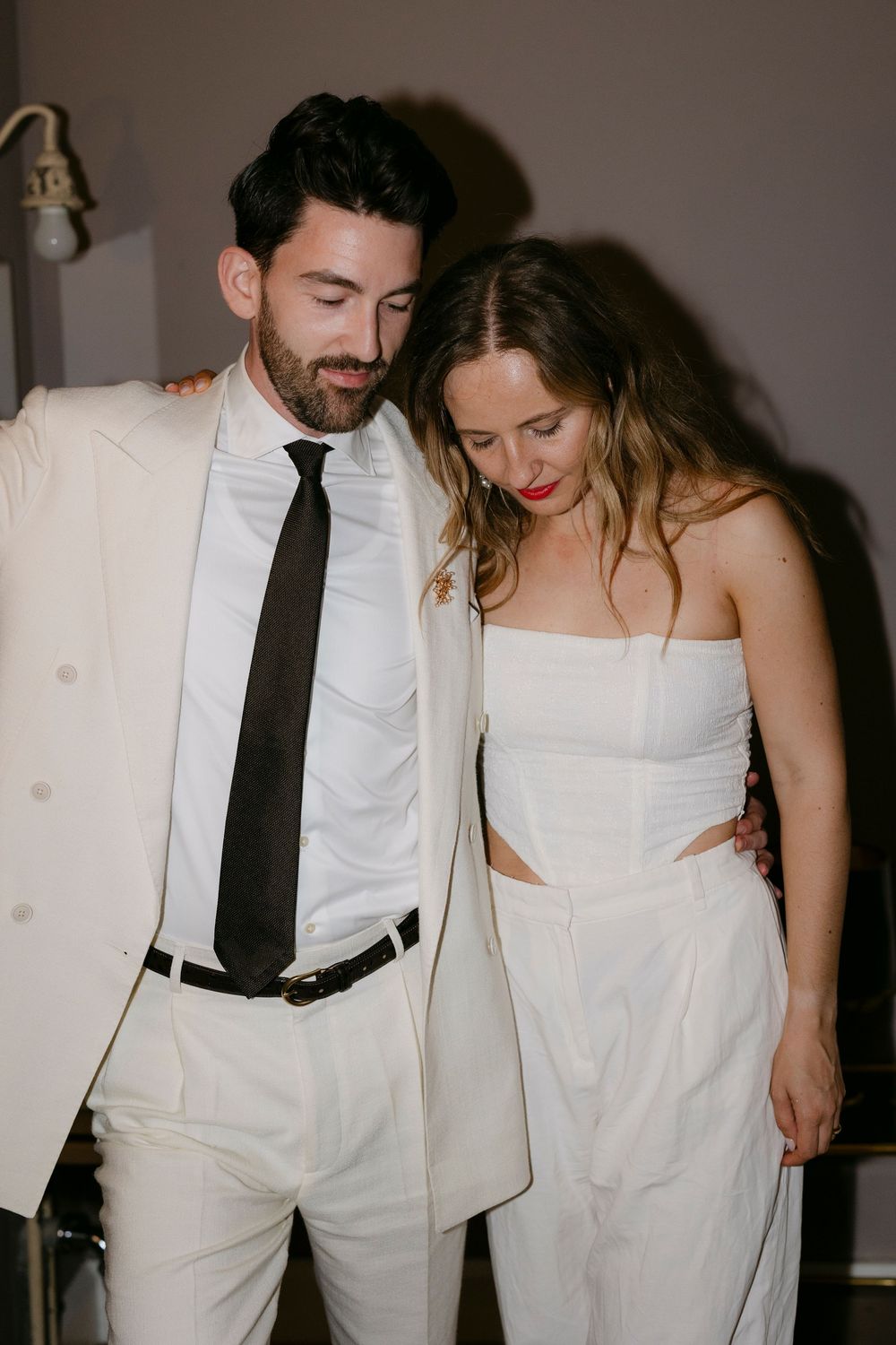 Couple in matching white outfits embrace closely, the man in a cream suit with a dark tie and the woman in a strapless top.