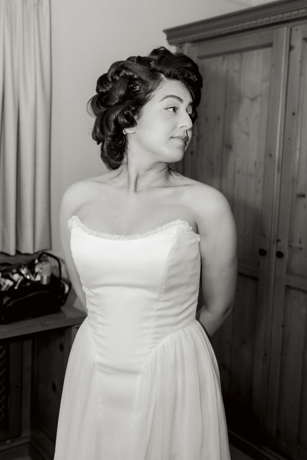 Black and white portrait of a bride in a strapless gown with pinned-up curls, glancing over her shoulder beside a wooden wardrobe.