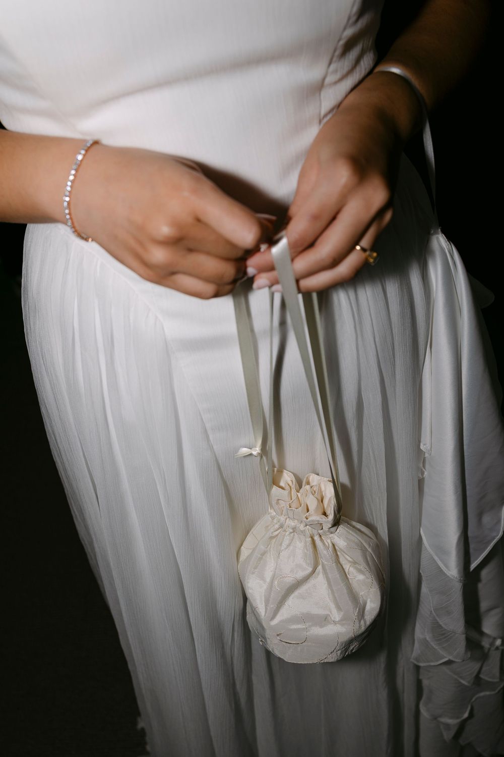 Bride in a white chiffon wedding gown holding a small ivory silk pouch purse by its ribbon straps, wearing a bracelet and ring.