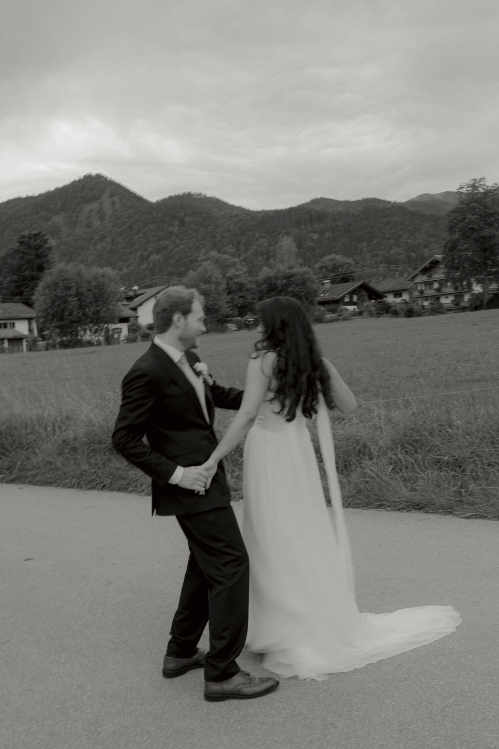 Black and white photo of a bride and groom holding hands on a country road with a grassy meadow and forested mountains behind them.