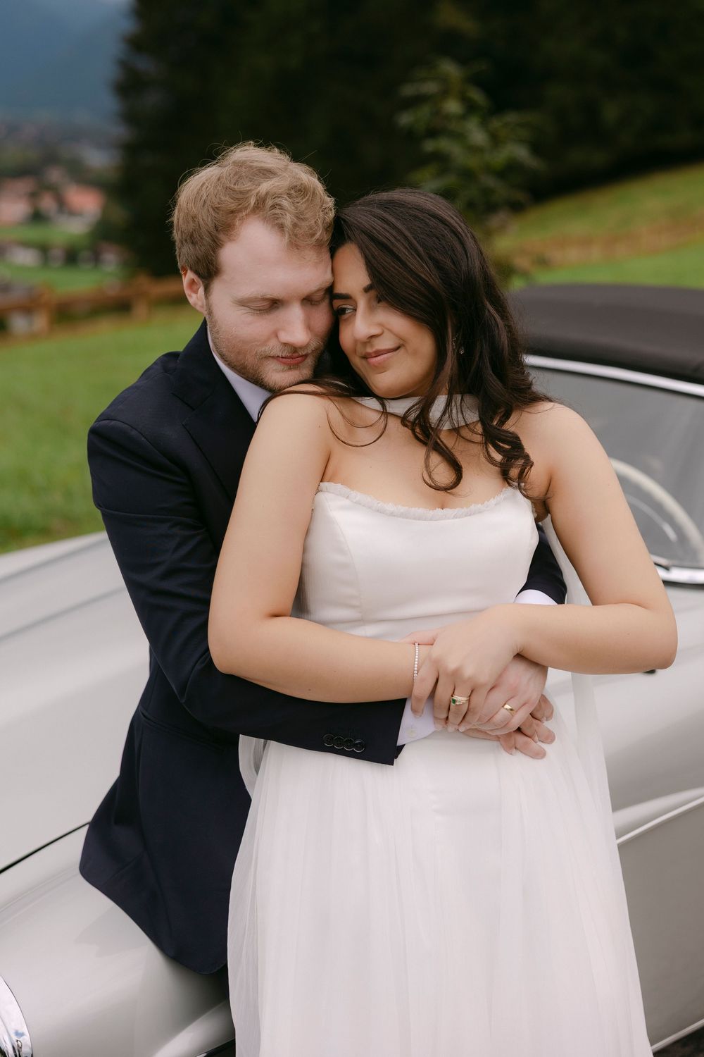 Bride in a white strapless gown embraced from behind by a groom in a navy suit beside a vintage silver car with green hills behind.