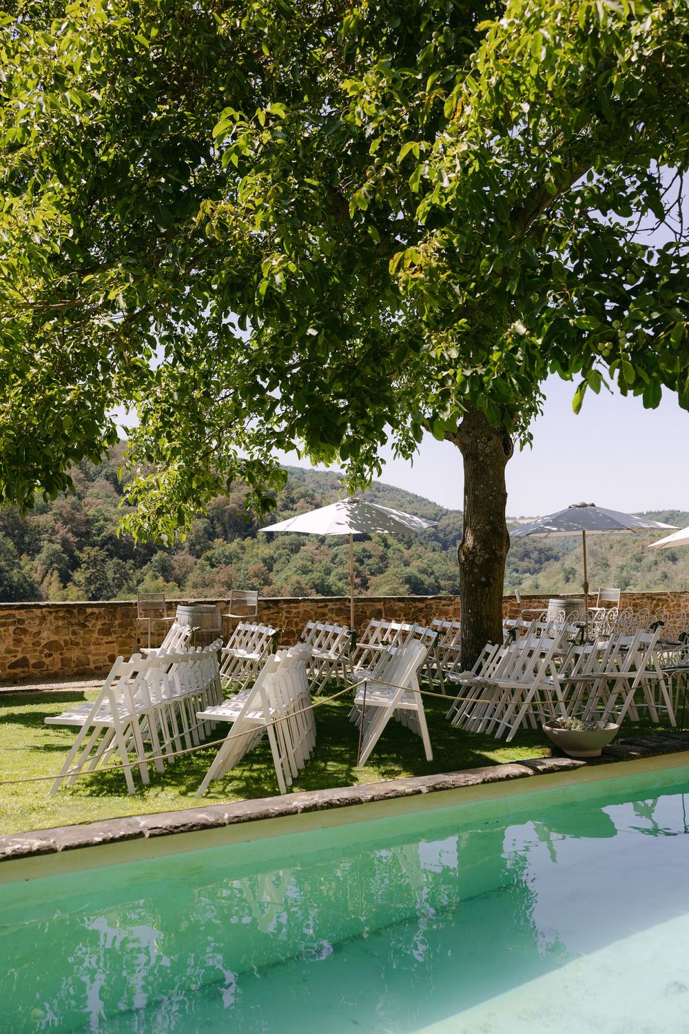 White folding chairs set up under a shady tree beside a turquoise swimming pool, with umbrellas and rolling hills beyond a stone wall.