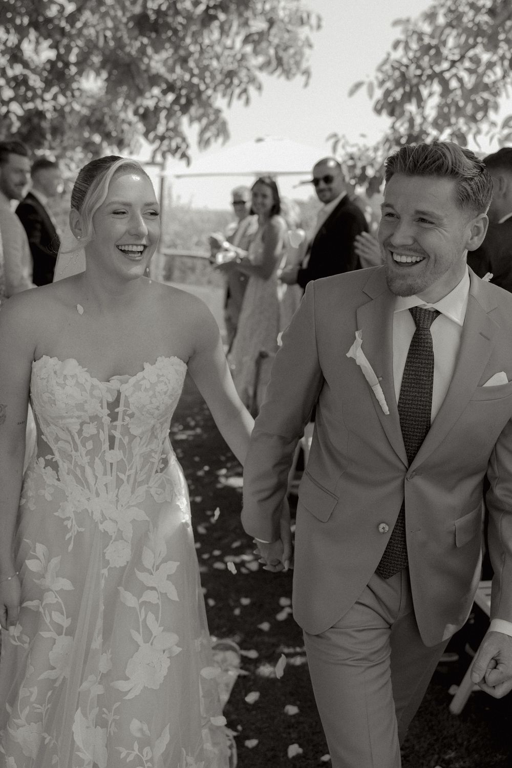 Smiling bride in a lace strapless gown holds hands with a laughing groom in a suit and tie as wedding guests gather behind them outdoors.
