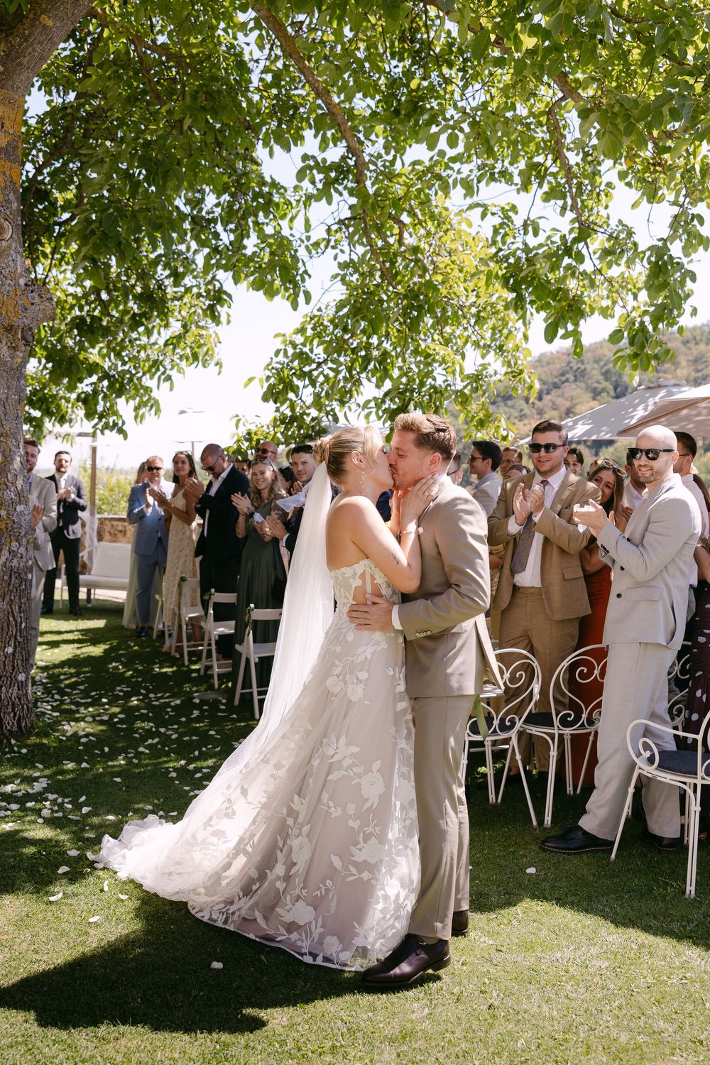 Bride in a lace wedding gown kisses her groom in a tan suit as guests applaud during an outdoor ceremony under a tree.