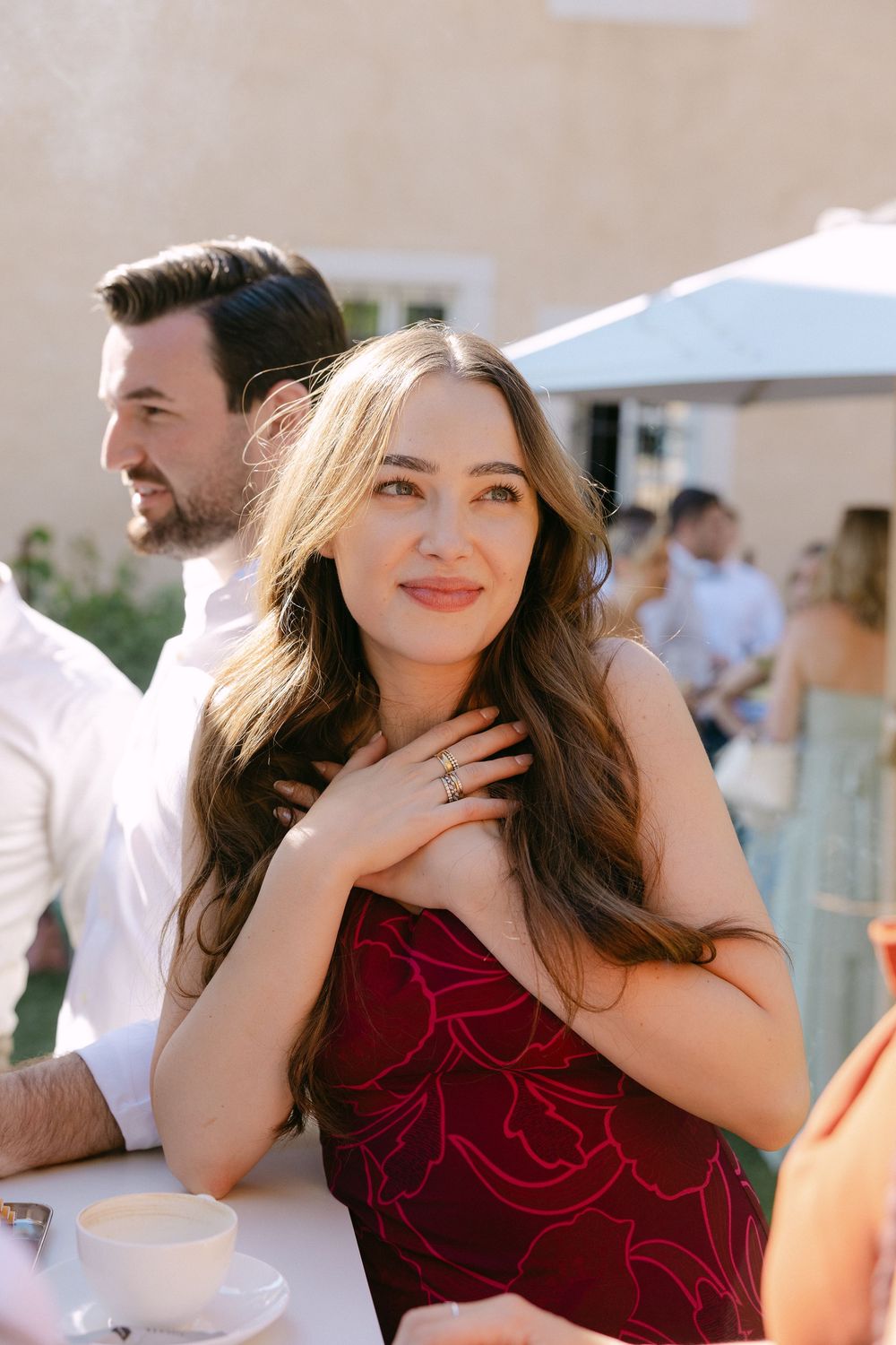 Smiling woman in a burgundy satin dress with long wavy hair clasps her hands at her chest at a sunny outdoor garden party.