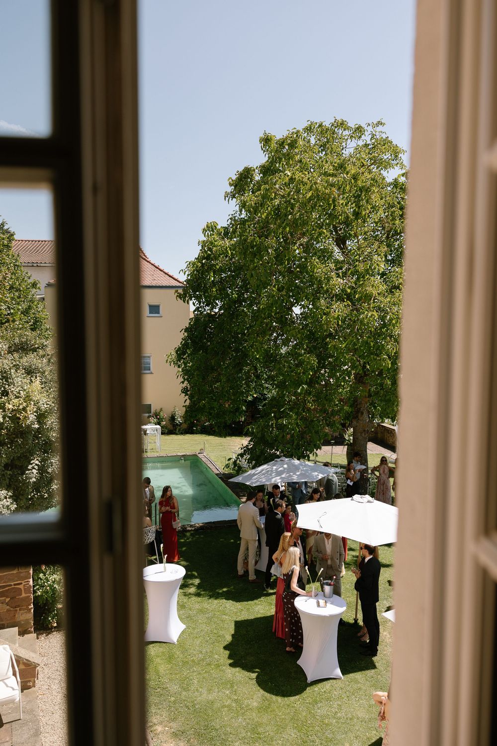View through an open window of guests mingling at an outdoor garden party with cocktail tables, white umbrellas, a pool and a large tree.