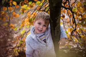 Cute smiling child wearing a light grey fleece hugging a tree and surrounded by autumnal leaves